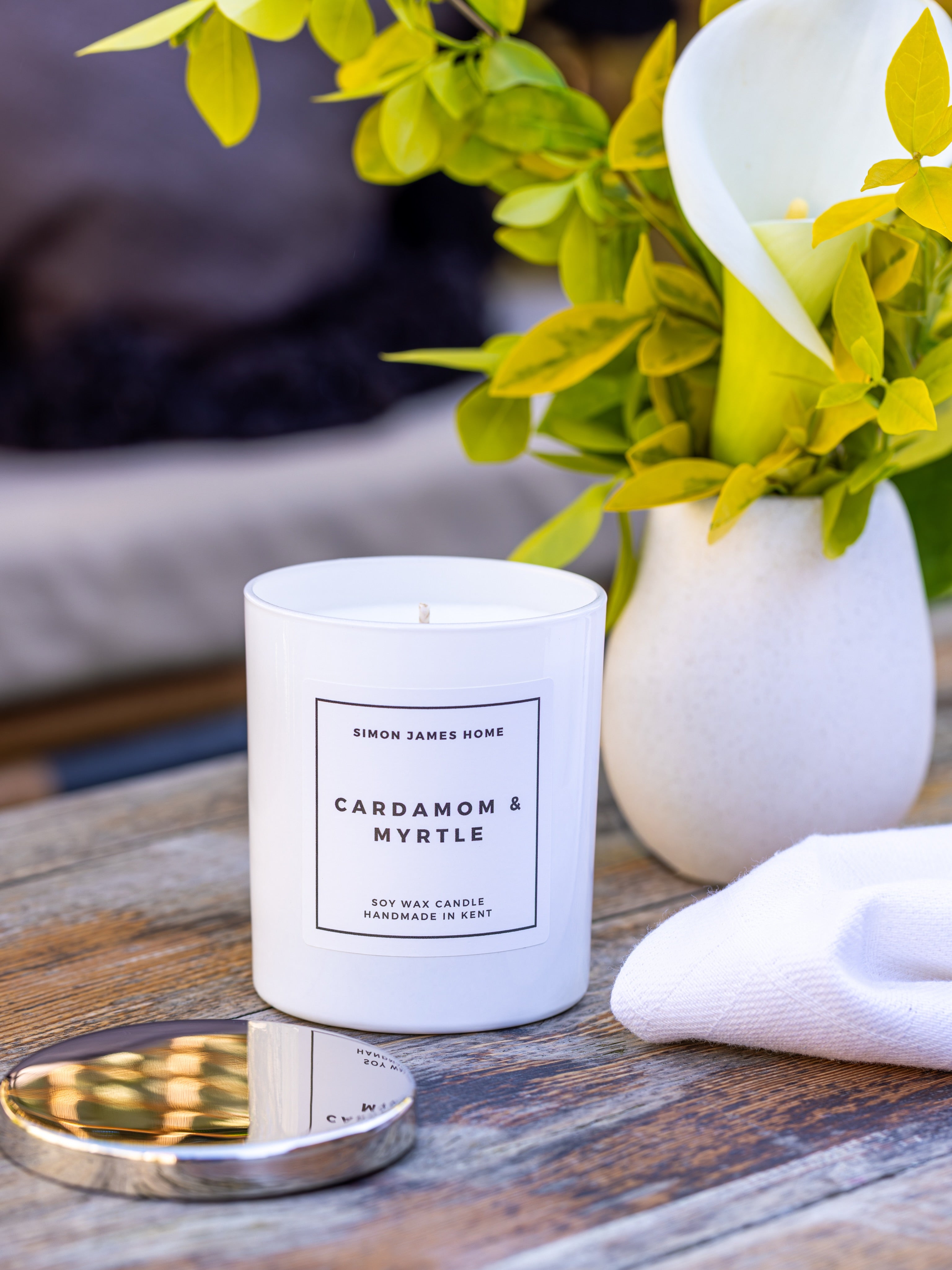 A white soy candle jar labeled 'CARDAMOM & MYRTLE' by Simon James Home sits on a wooden table beside a reflective silver lid, with a white vase of green leaves and a folded white cloth in the background.