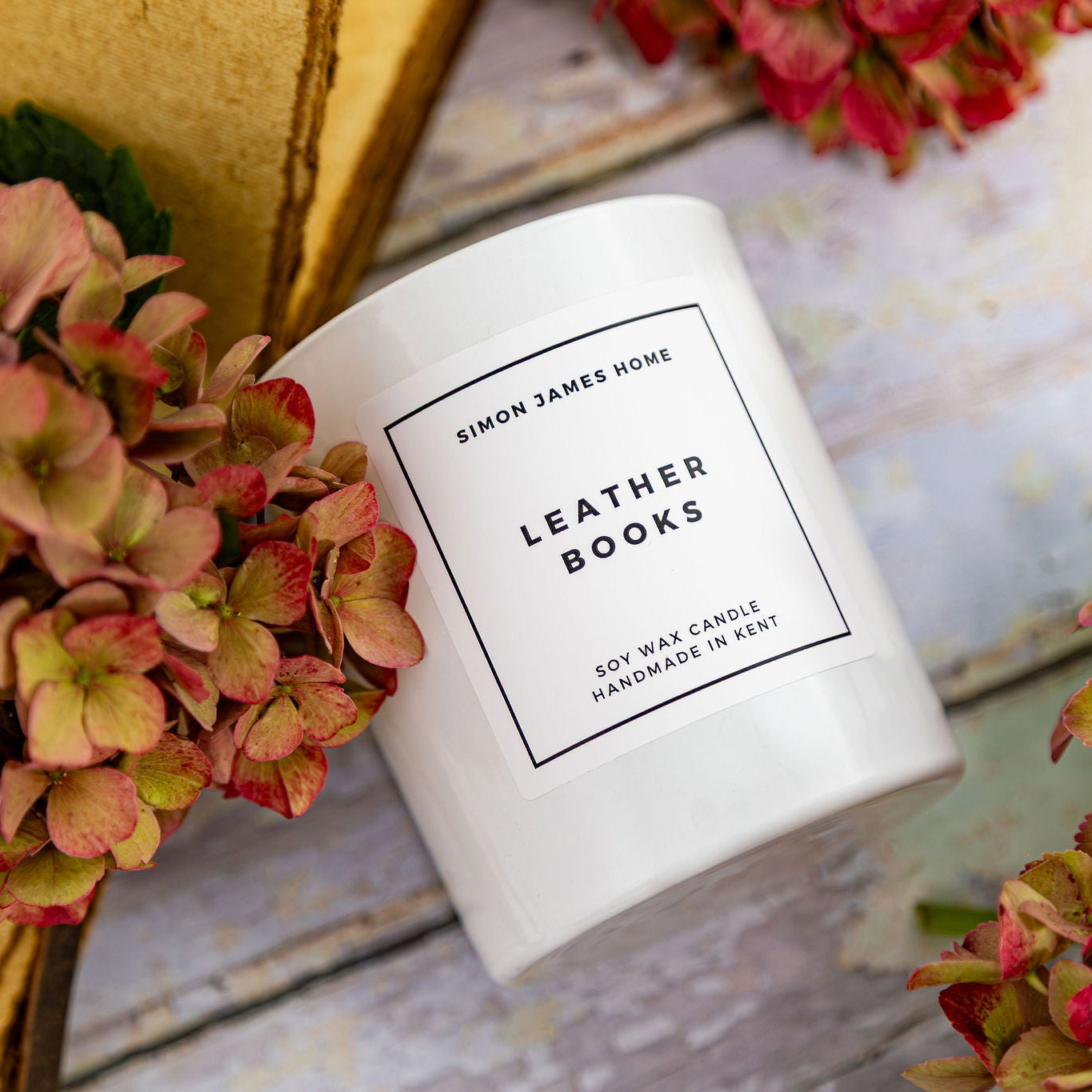 A white soy wax candle jar labeled 'Leather Books' by Simon James Home, styled with dried pink hydrangeas and a gold-toned book on a rustic whitewashed wooden surface.