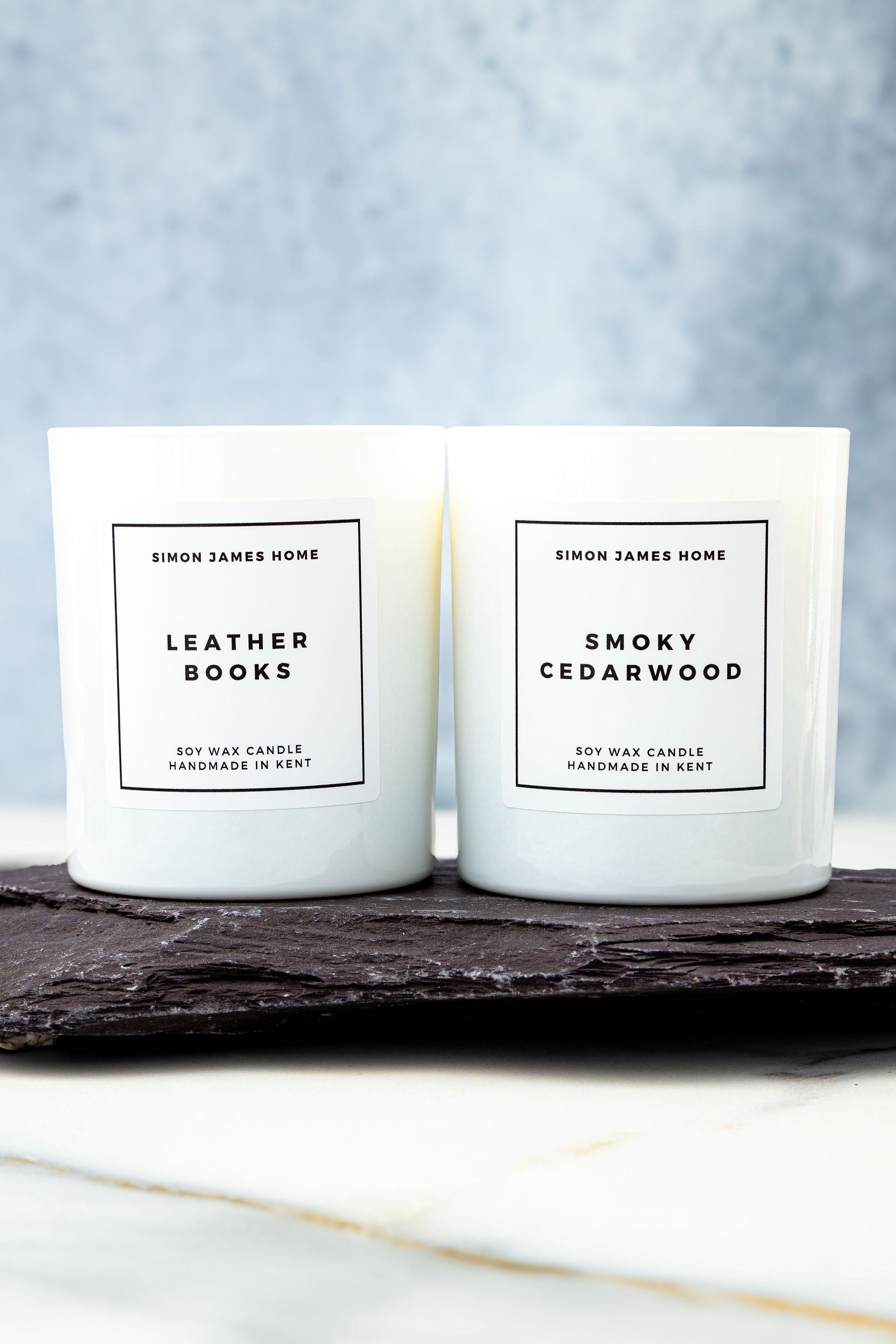 Two candles labeled 'Leather Books' and 'Smoky Cedarwood' by Simon James Home on a dark surface with a light background.