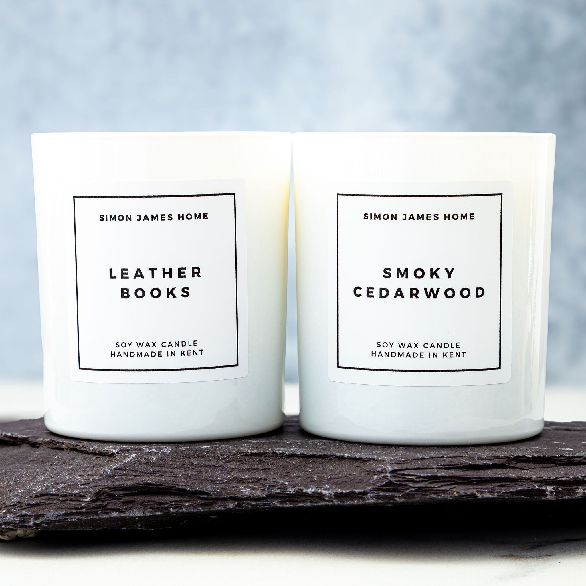 Two candles labeled 'Leather Books' and 'Smoky Cedarwood' by Simon James Home on a dark surface with a light background.