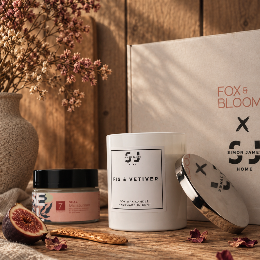 A styled product shot featuring a white wax candle jar labeled 'FIG & VETIVER' alongside its metallic lid, a small jar with a black lid, and a branded box reading 'FOX & BLOOM' and 'SIMON JAMES HOME', arranged on a rustic wooden surface with dried flowers, a fig, and a burlap cloth.