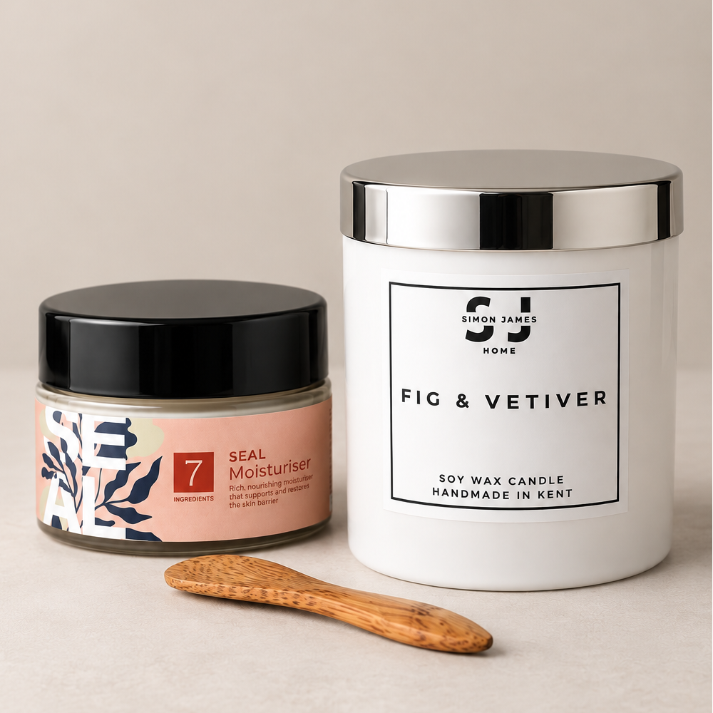 A styled product shot featuring a white soy wax candle jar with a silver lid labeled 'Fig & Vetiver' by 'Simon James Home', a smaller peach-coloured jar of 'Seal Moisturizer' from Fox & Bloom with a black lid, and a small wooden spoon on a neutral beige background.