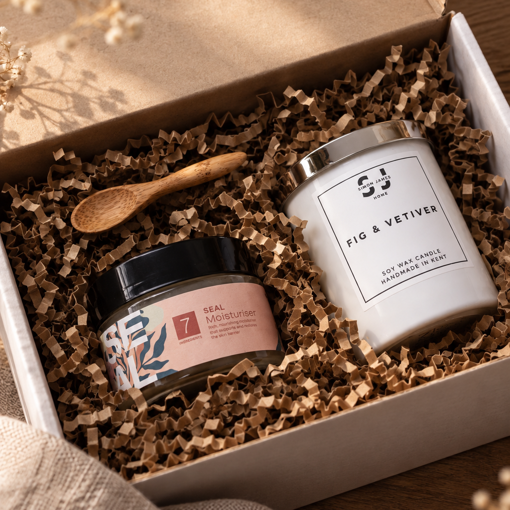 A gift box set containing a white jar candle labeled 'FIG & VETIVER' and a smaller pink-labeled jar of natural moisturiser, nestled in brown crinkle paper inside an open kraft box with a small wooden spoon.