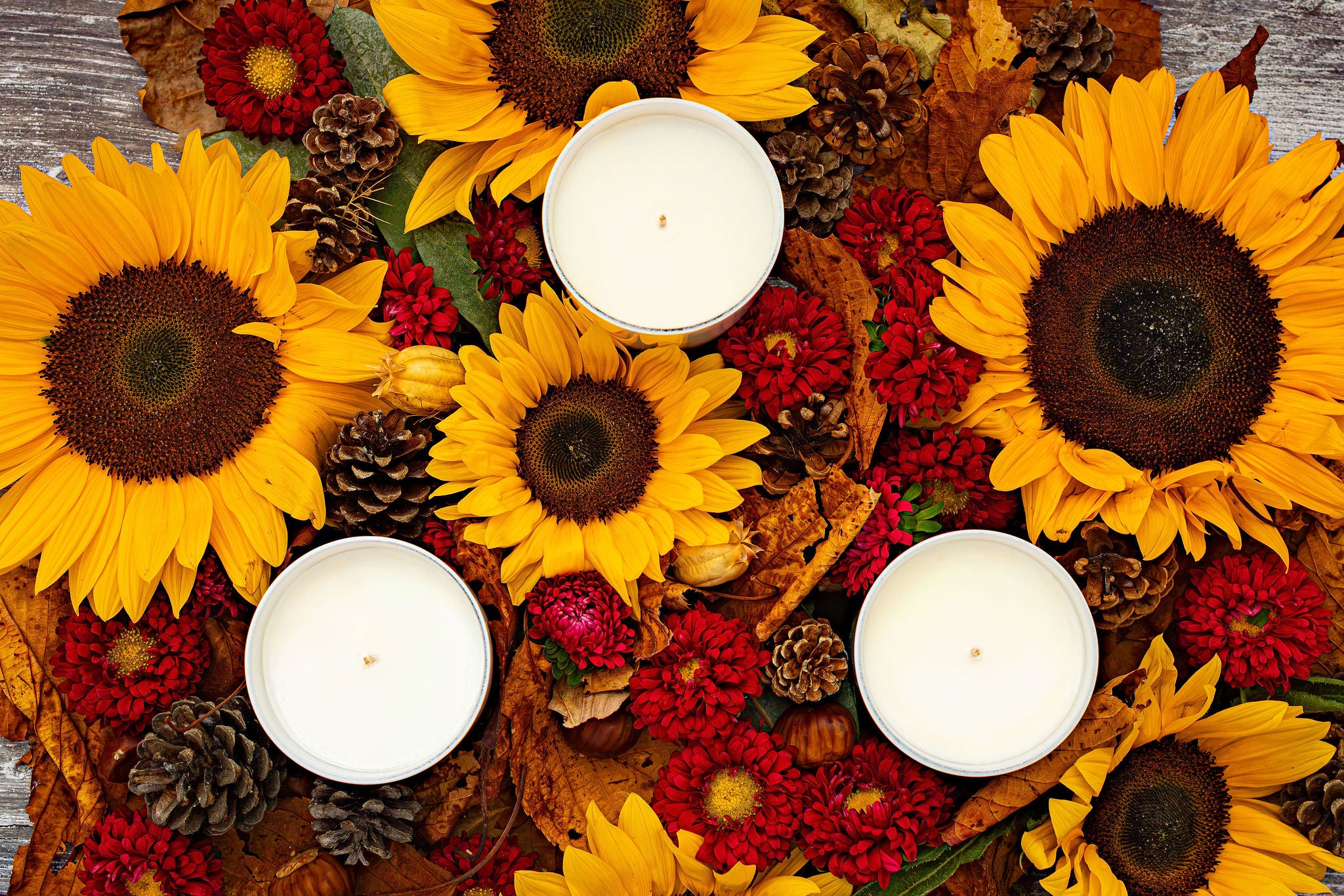 Three white pillar candles arranged in a rustic autumn floral arrangement with sunflowers, red flowers, pinecones, and dried leaves on a weathered wood surface.