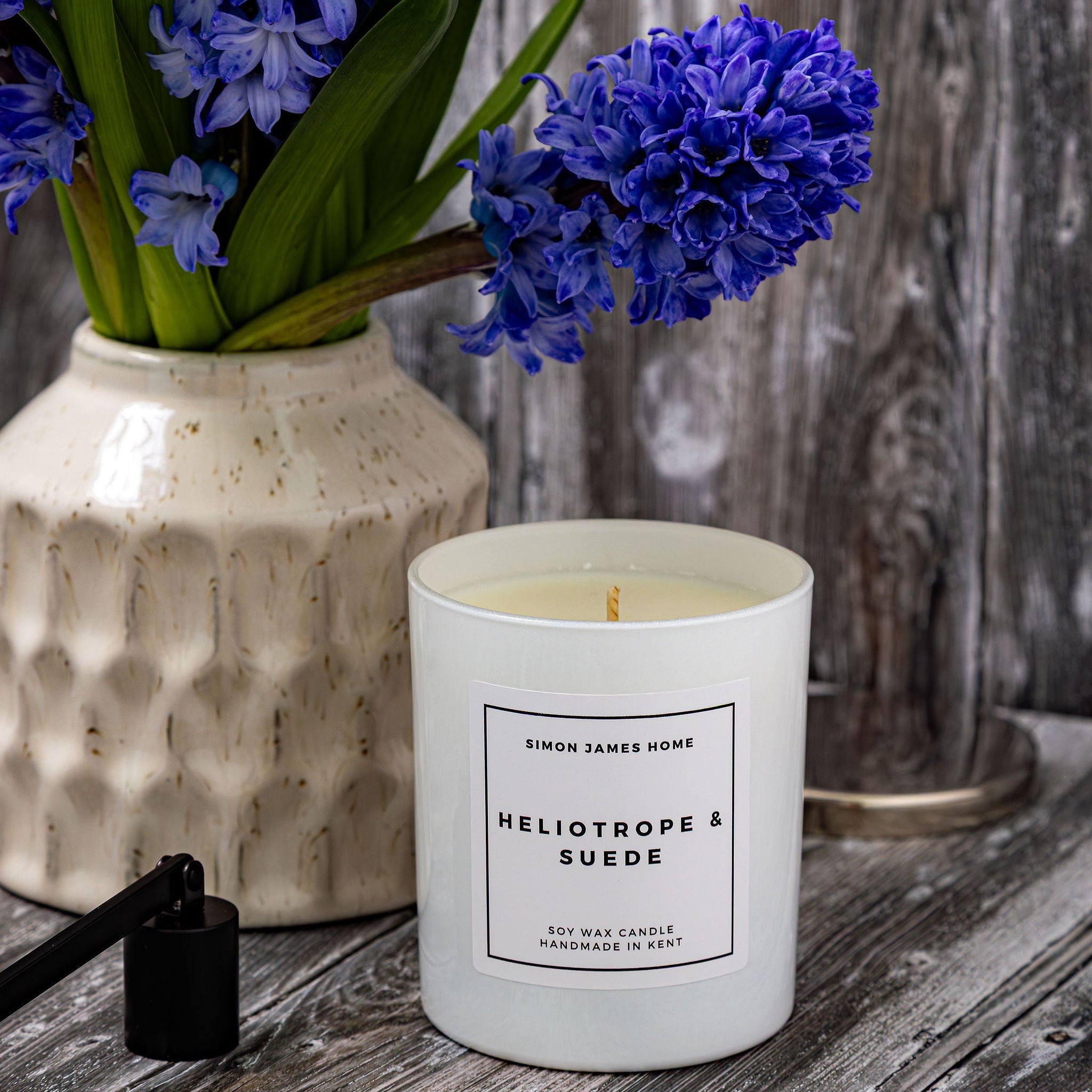 Candle labeled 'Heliotrope & Suede' with blue flowers in a vase on a wooden surface