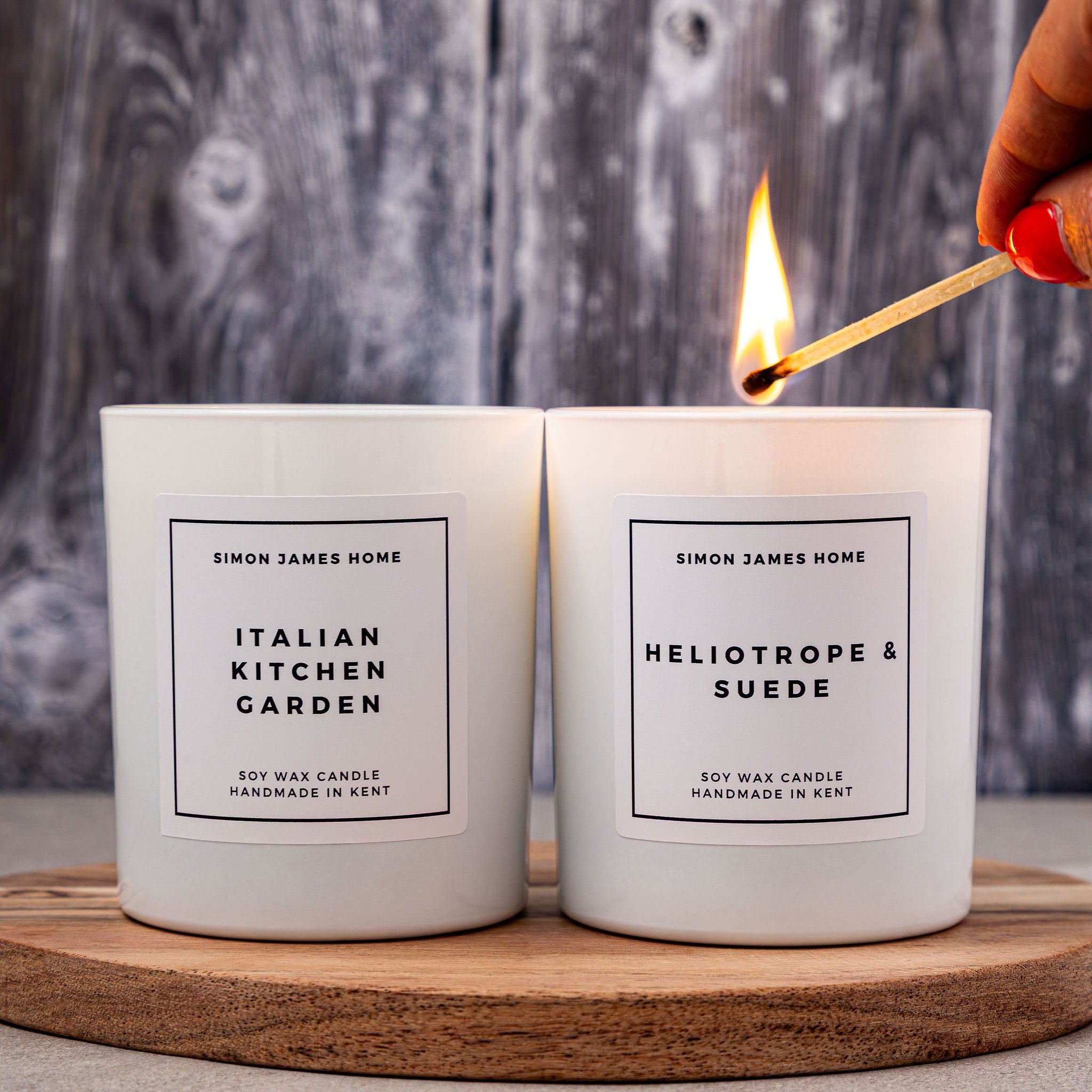 Two candles labeled 'Italian Kitchen Garden' and 'Heliotrope & Suede' on a wooden board with a match igniting one of them.