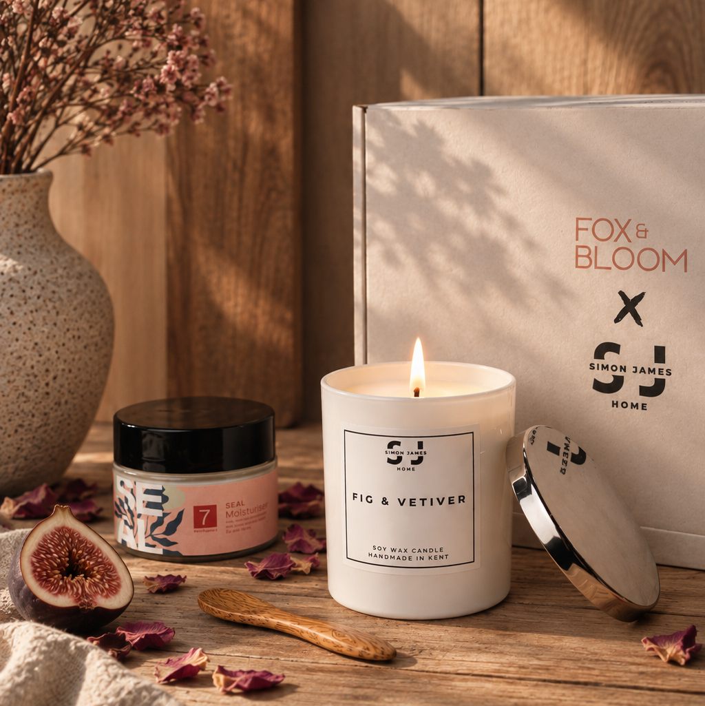 A lit white candle labeled 'Simon James Home FIG & VETIVER' sits on a rustic wooden table beside its white gift box, with a jar, dried flowers in a vase, and scattered rose petals in a warm styled setting.