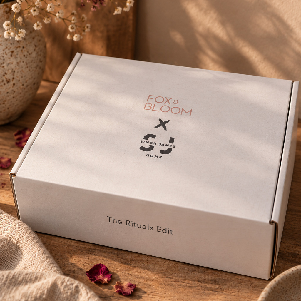 A white subscription box labeled 'FOX + BLOOM x Simon James Home Rituals Edit' sits on a wooden surface with dried flowers, scattered rose petals, and soft warm shadows in the background.